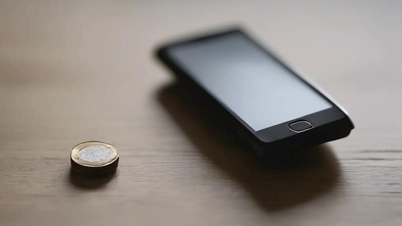 Smartphone mit dunklem, ausgeschaltetem Display liegt auf einem Holztisch neben einer einzelnen Münze